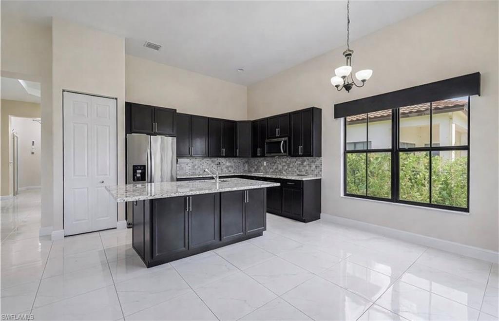 Kitchen featuring dark cabinets, stainless steel appliances, an island with sink, tasteful backsplash, and light stone countertops