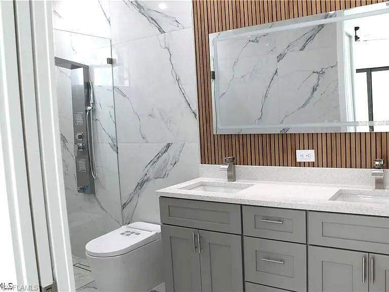 Bathroom featuring a dual vanity with light gray cabinetry and white speckled countertops, a large illuminated mirror, a wood-slat accent wall, and a walk-in shower with large format marble-look tile
