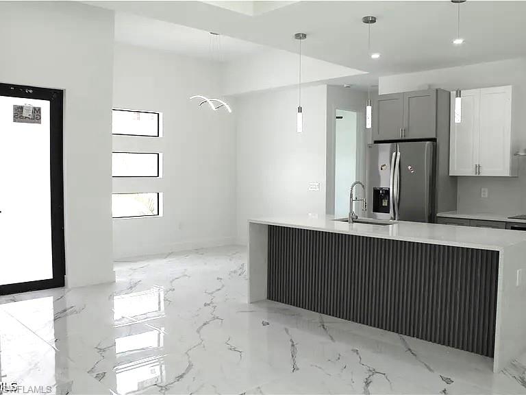 Open-concept kitchen and living area featuring white tile flooring, a kitchen island with fluted paneling and a waterfall countertop, stainless steel appliances, and contemporary pendant lighting