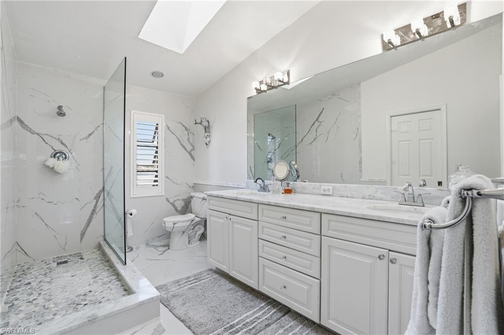 Primary bath featuring a skylight, double vanity, plantation shutters, marble finished shower, and light marble flooring