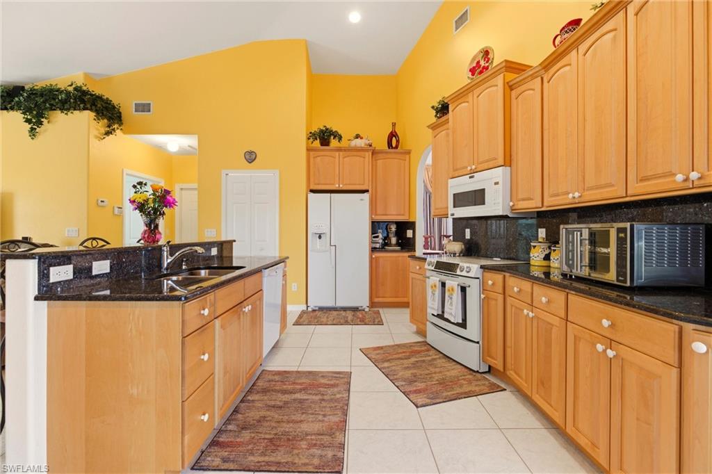 Kitchen featuring white appliances, dark stone countertops, light tile patterned flooring, a kitchen island with sink, and lofted ceiling