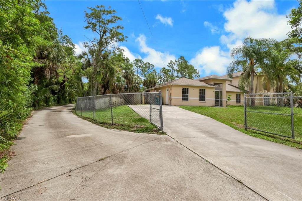 Gated concrete driveway leading to a stucco exterior residence with multiple windows, surrounded by mature trees and green landscaping