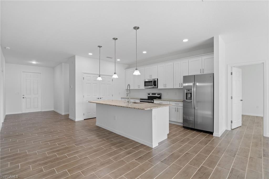 Kitchen featuring stainless steel appliances, pendant lighting, an island with sink, light stone countertops, and white cabinetry