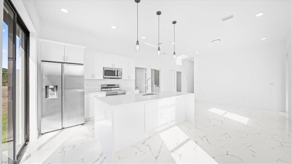 Kitchen featuring stainless steel appliances, white cabinets, decorative light fixtures, a kitchen island with sink, and light stone countertops