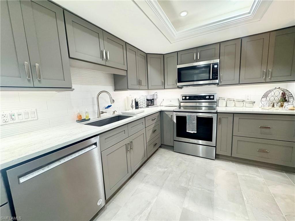 Kitchen featuring gray cabinetry, stainless steel appliances, light stone counters, crown molding, and a raised ceiling