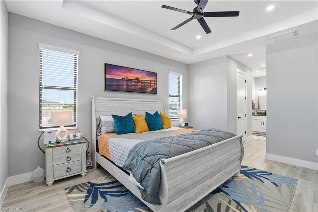 Bedroom with a raised ceiling, light wood-style floors, ensuite bath, ceiling fan, and recessed lighting