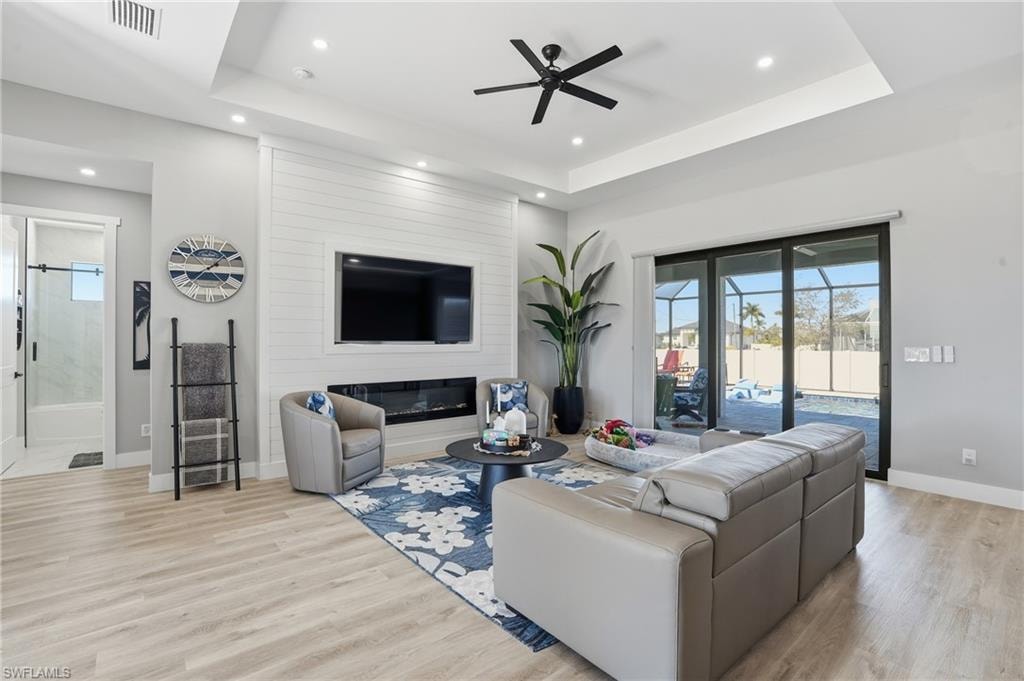 Living room featuring a raised ceiling, ceiling fan, light wood-type flooring, a large fireplace, and recessed lighting