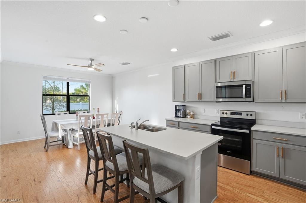 Kitchen featuring gray cabinetry, stainless steel appliances, crown molding, a center island with sink, and light wood-type flooring