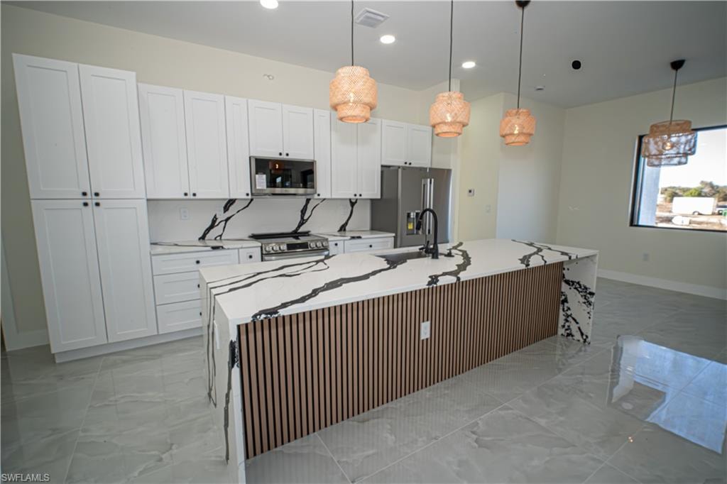 Kitchen with light stone counters, stainless steel appliances, a center island with sink, hanging light fixtures, and light marble finish floors