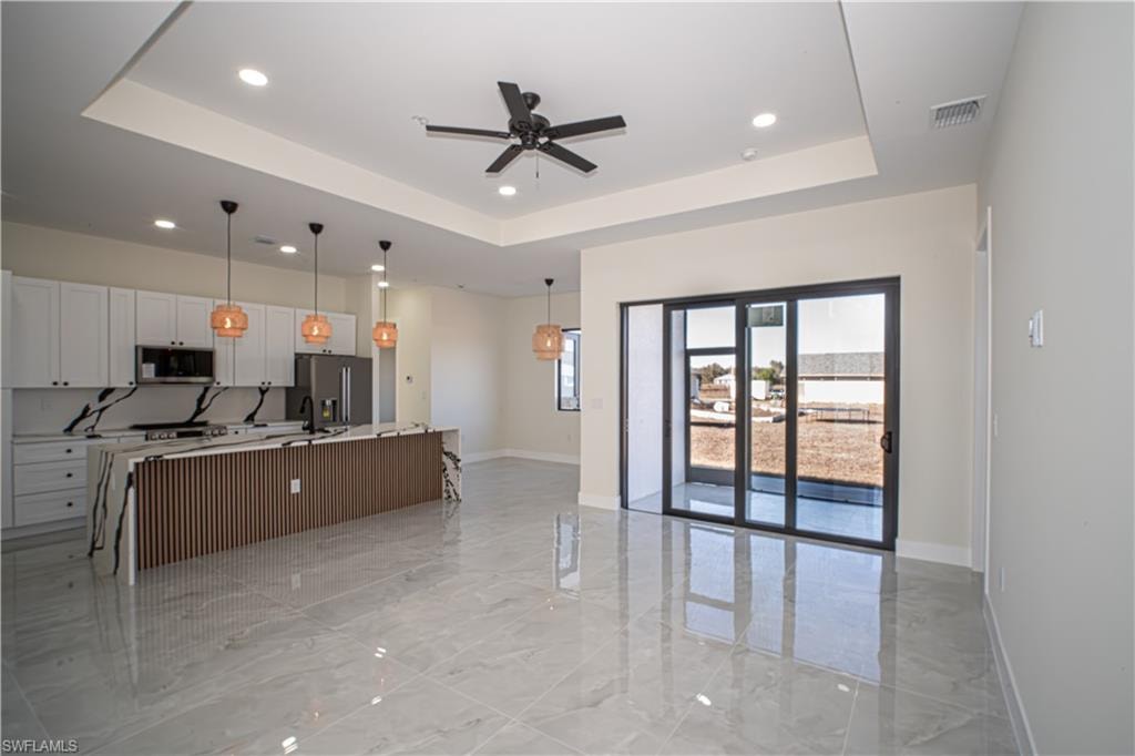Kitchen with white cabinets, a center island with sink, hanging light fixtures, light marble finish flooring, and a tray ceiling