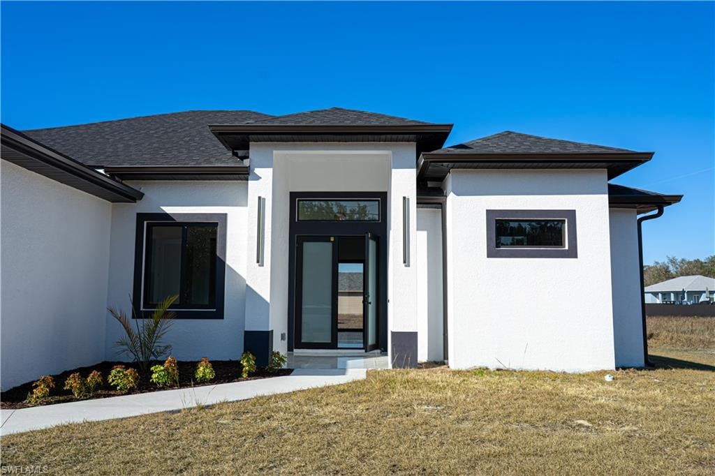 Doorway to property featuring a yard, stucco siding, and a shingled roof