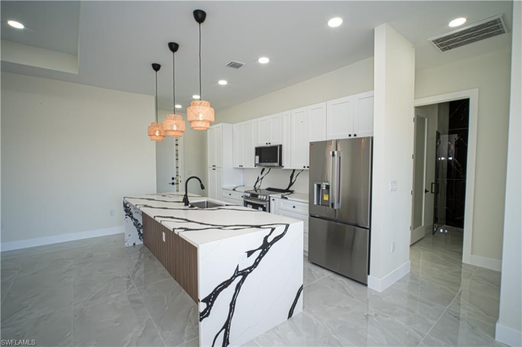 Kitchen with stainless steel appliances, an island with sink, white cabinetry, pendant lighting, and light marble finish floors