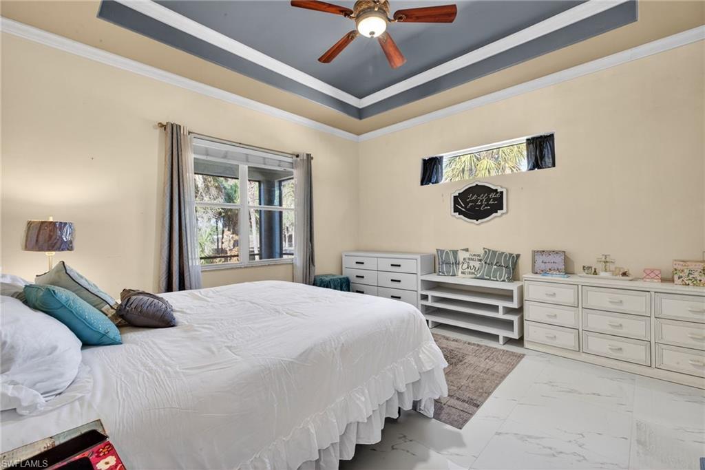 Bedroom featuring light marble finish floors, multiple windows, a raised ceiling, a ceiling fan, and ornamental molding