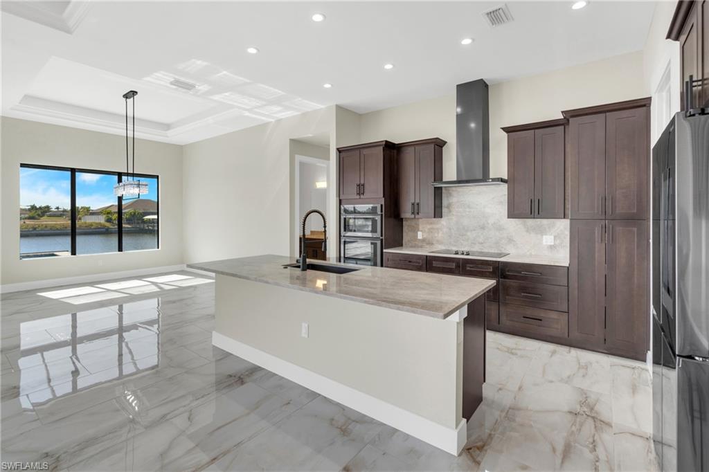 Kitchen with light stone countertops, a kitchen island with sink, stainless steel appliances, light marble finish floors, and range hood