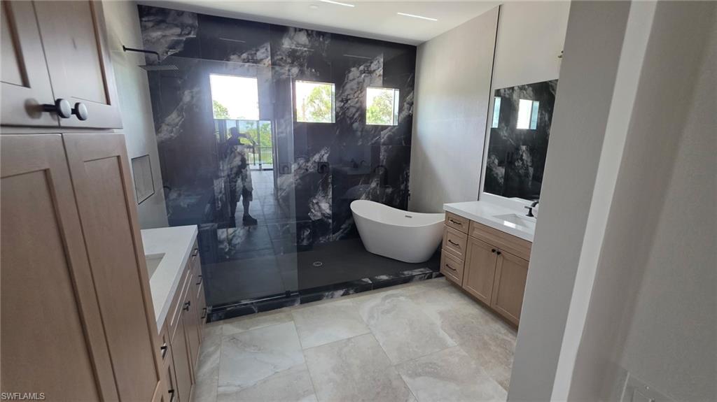 Freestanding soaking tub, dual vanity with light wood cabinetry, walk-in shower with black-veined tile and glass enclosure, and light-toned floor tile