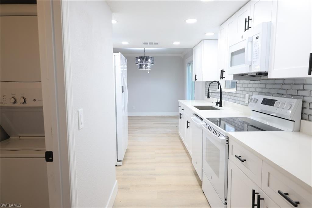 Kitchen with white appliances, white cabinetry, light wood finished floors, stacked washing machine and dryer, and backsplash
