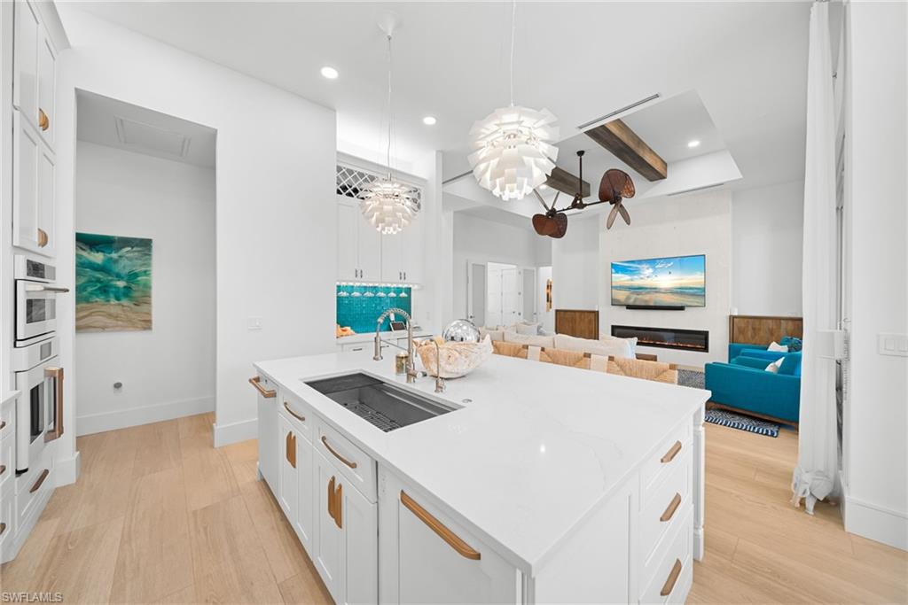 Kitchen featuring white cabinets, beam ceiling, light stone counters, an island with sink, and decorative light fixtures