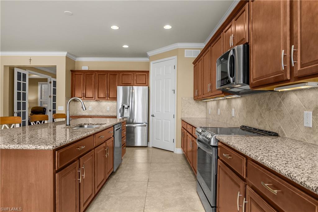 Kitchen featuring stainless steel appliances, wood finish cabinets, light stone countertops, ornamental molding, and decorative backsplash