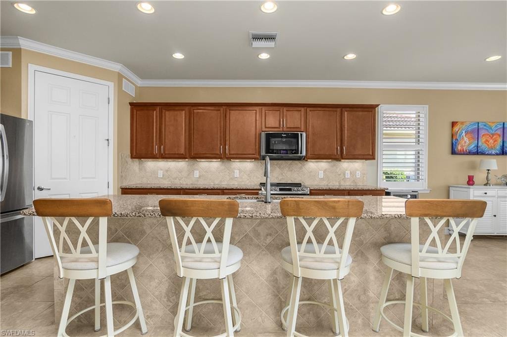 Kitchen featuring stainless steel appliances, wood finish cabinetry, light stone countertops, a kitchen breakfast bar, and a center island with sink