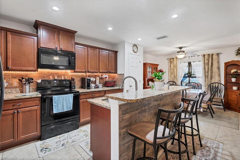 Kitchen featuring black appliances, a ceiling fan, a kitchen island with sink, backsplash, and light stone countertops