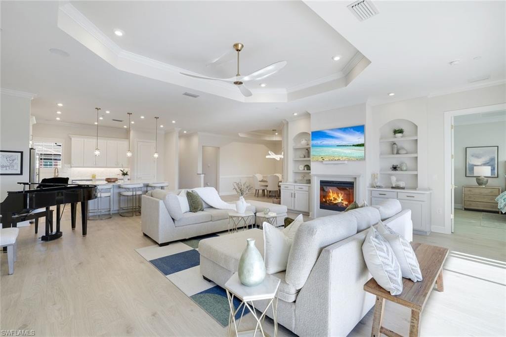 Living area with built in shelves, light wood-type flooring, ornamental molding, ceiling fan, and a tray ceiling