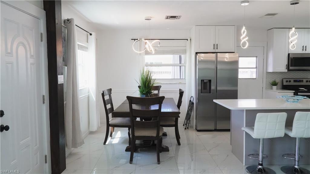 Kitchen featuring stainless steel appliances, a kitchen bar, white cabinets, hanging light fixtures, and light marble finish flooring