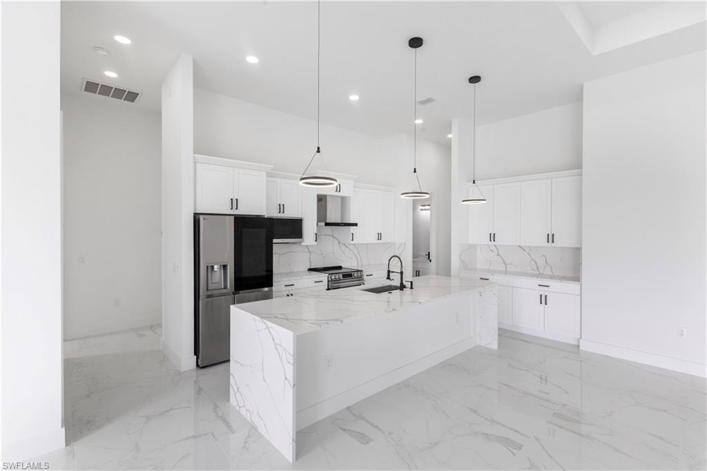 Kitchen featuring stainless steel appliances, light stone countertops, a kitchen island with sink, white cabinetry, and hanging light fixtures