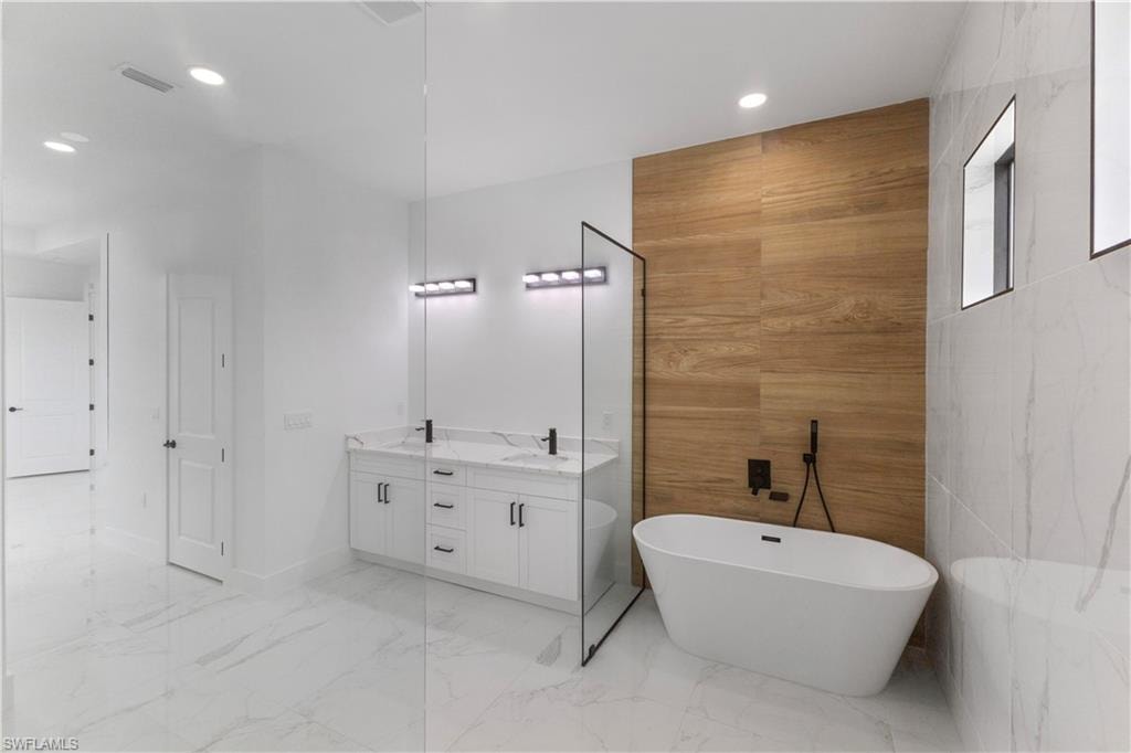 Bathroom featuring double vanity, tile walls, a freestanding tub, light marble finish flooring, and recessed lighting