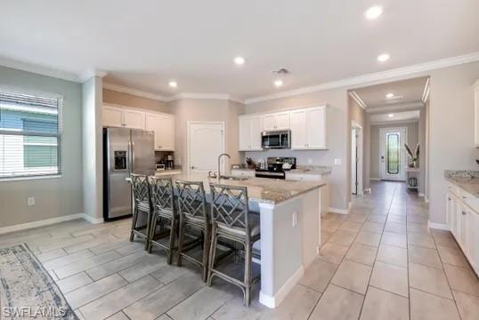 Kitchen with white cabinets, stainless steel appliances, a kitchen breakfast bar, an island with sink, and light stone counters