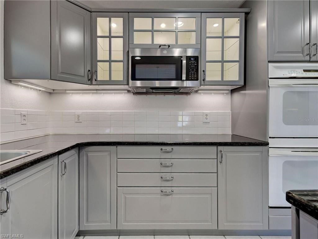 Kitchen featuring white double oven, stainless steel microwave, glass insert cabinets, dark stone counters, and gray cabinetry