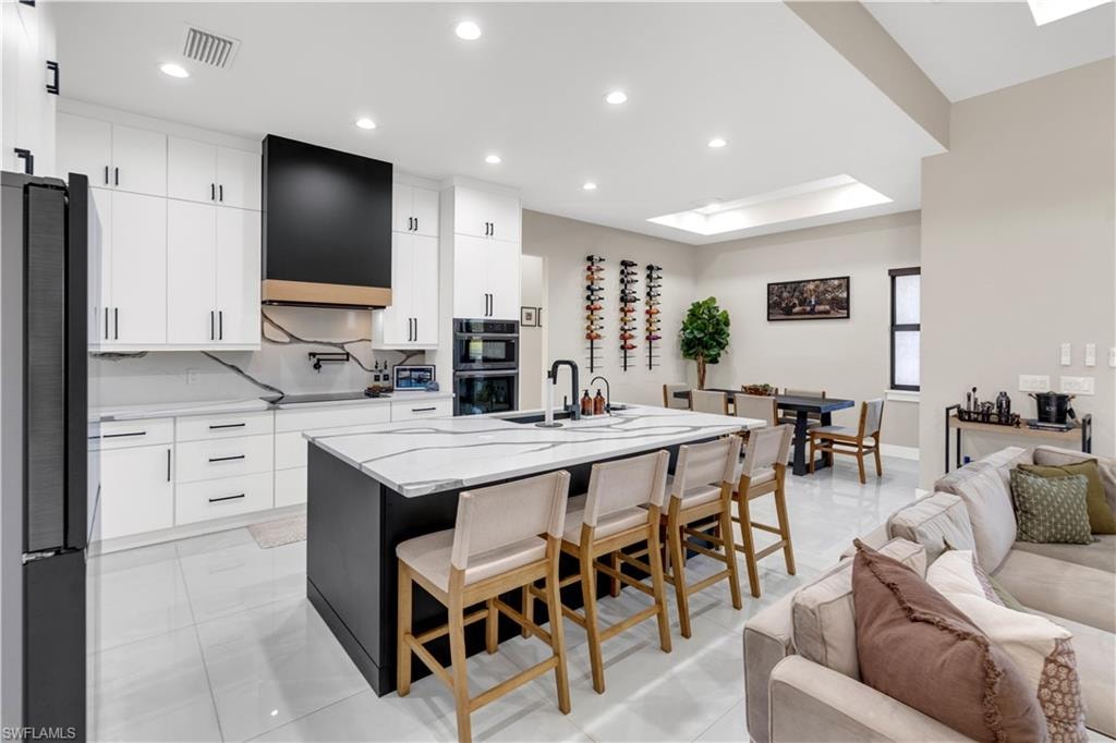 Two tone kitchen with a kitchen breakfast bar, light stone countertops, a center island with sink, black appliances, and modern cabinets