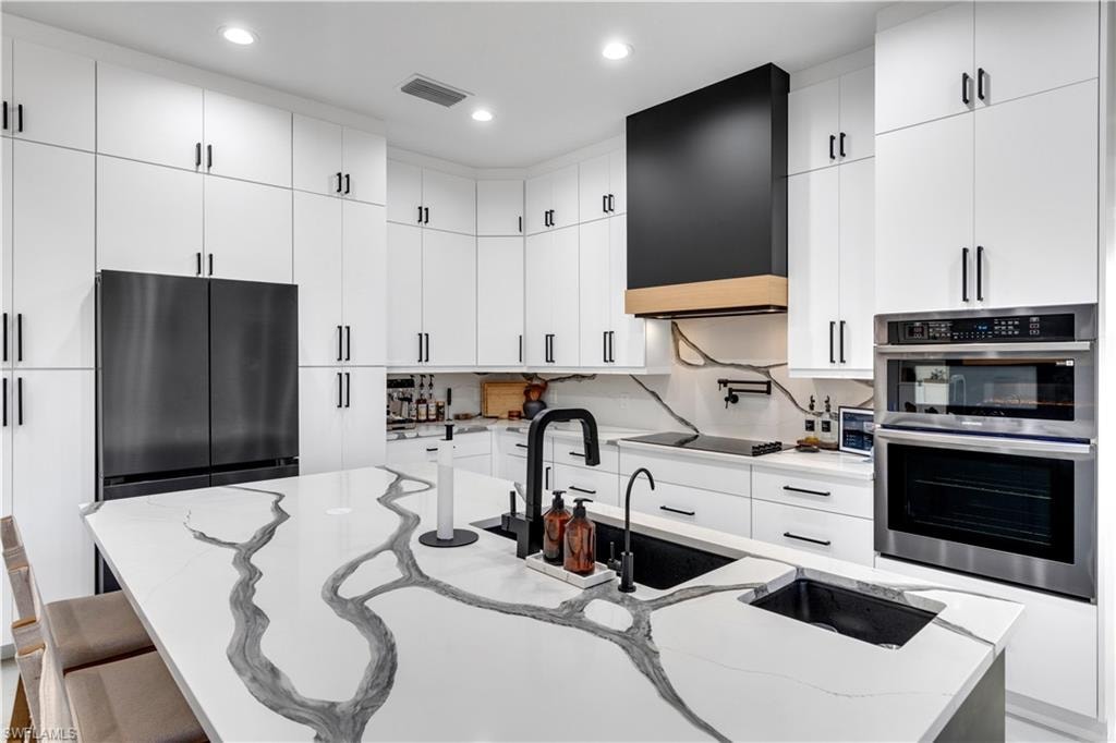 Kitchen with stainless steel appliances, a kitchen island with sink, light stone countertops, white cabinetry, and recessed lighting