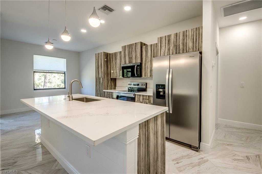 Kitchen featuring wood finish cabinetry, stainless steel appliances, decorative light fixtures, a center island with sink, and light stone countertops