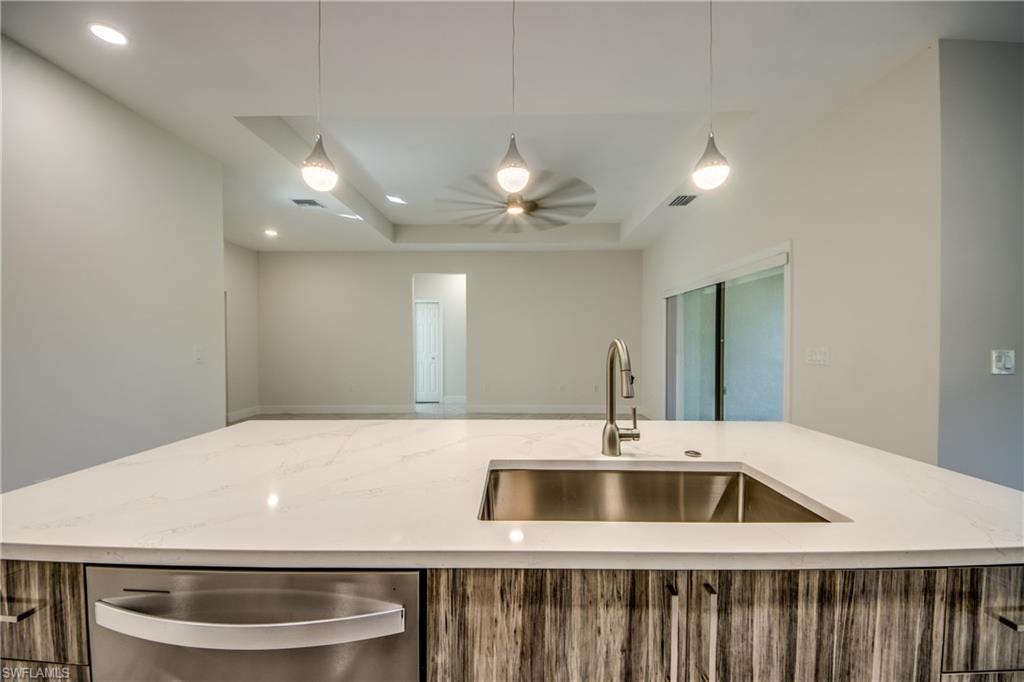Kitchen with light stone countertops, a raised ceiling, a center island with sink, stainless steel dishwasher, and pendant lighting
