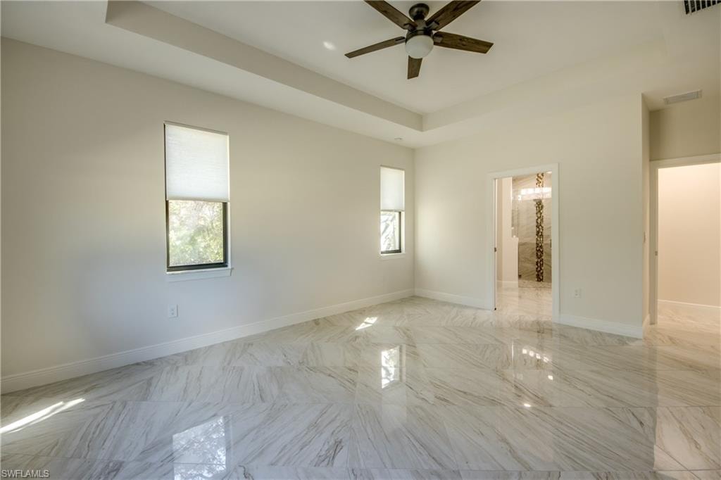 Unfurnished bedroom with a raised ceiling, multiple windows, a ceiling fan, ensuite bath, and marble finish floors