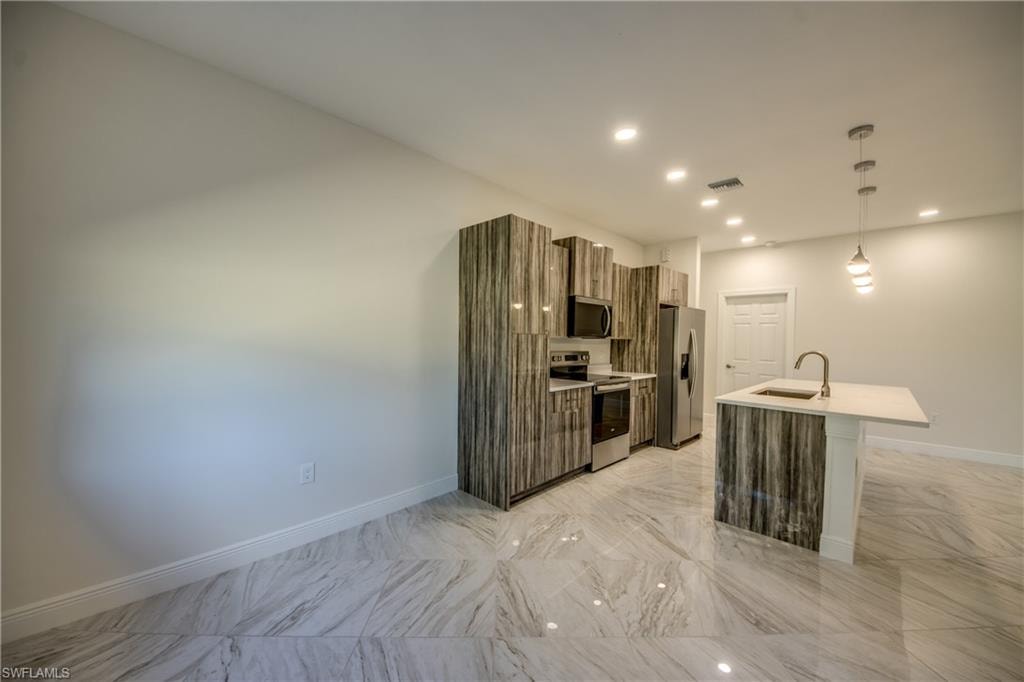 Kitchen featuring modern cabinets, stainless steel appliances, hanging light fixtures, a kitchen island with sink, and light stone countertops