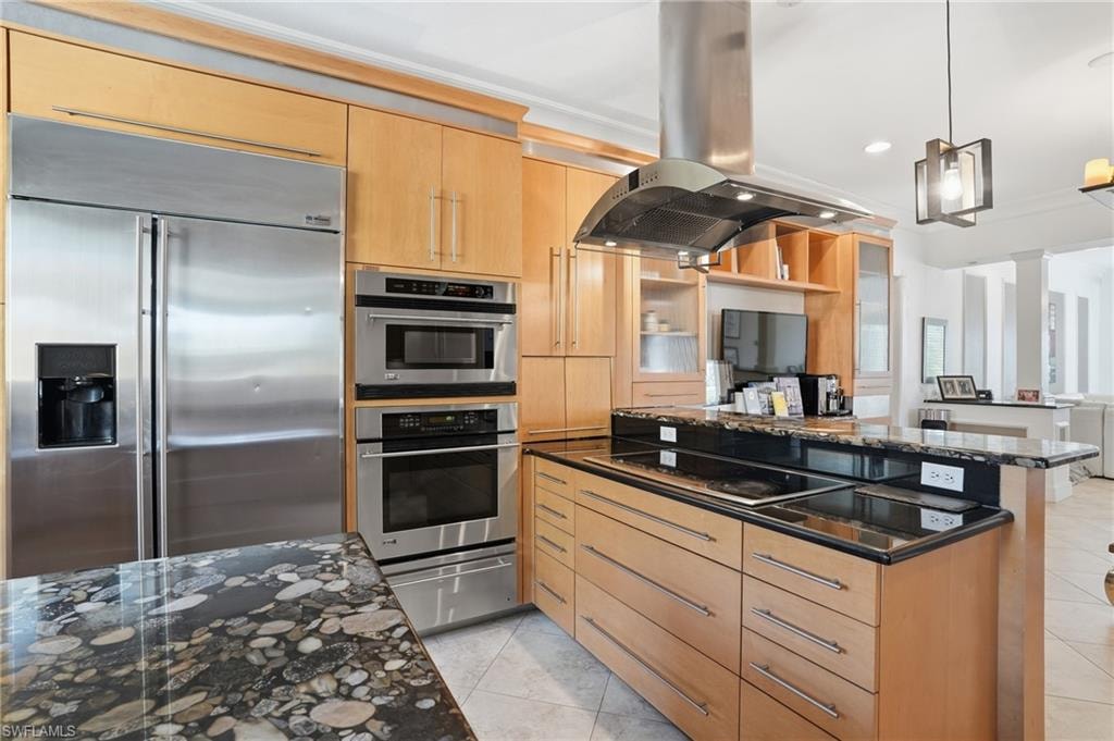 Kitchen with stainless steel appliances, island range hood, dark stone countertops, decorative light fixtures, and light wood finish cabinetry