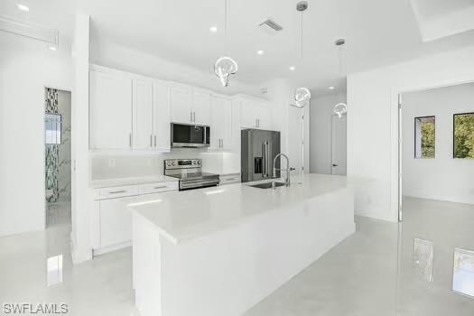 Kitchen with white cabinets, stainless steel appliances, an island with sink, hanging light fixtures, and concrete flooring