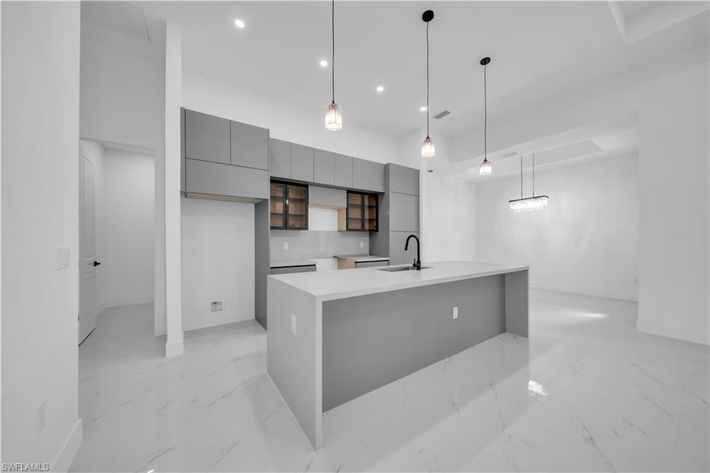 Kitchen featuring modern cabinets, gray cabinetry, hanging light fixtures, a center island with sink, and light stone counters