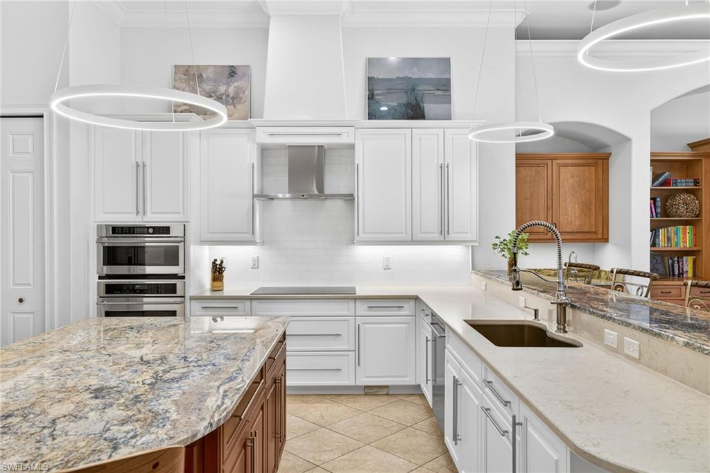 Kitchen with light stone countertops, ornamental molding, two tone cabinetry, decorative light fixtures, and stainless steel double oven