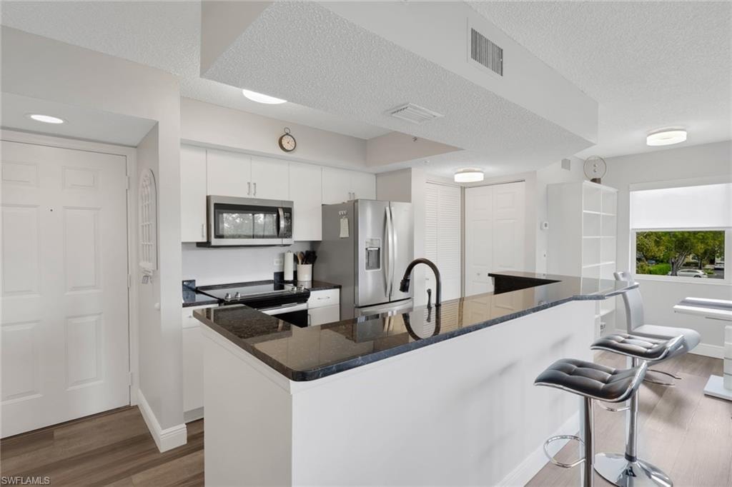 Kitchen featuring dark wood-style flooring, stainless steel appliances, white cabinets, dark stone countertops, and a breakfast bar area