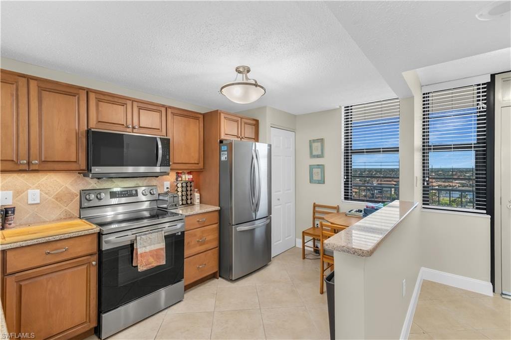 Kitchen with stainless steel appliances, wood finish cabinets, light stone counters, a textured ceiling, and light tile patterned floors