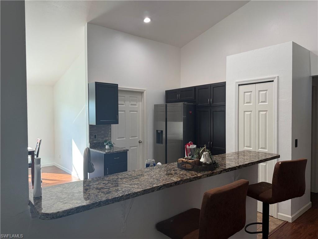 Kitchen view from living room featuring vaulted ceiling, dark wood-type flooring, a kitchen bar, stainless steel fridge with ice dispenser, and dark stone counters