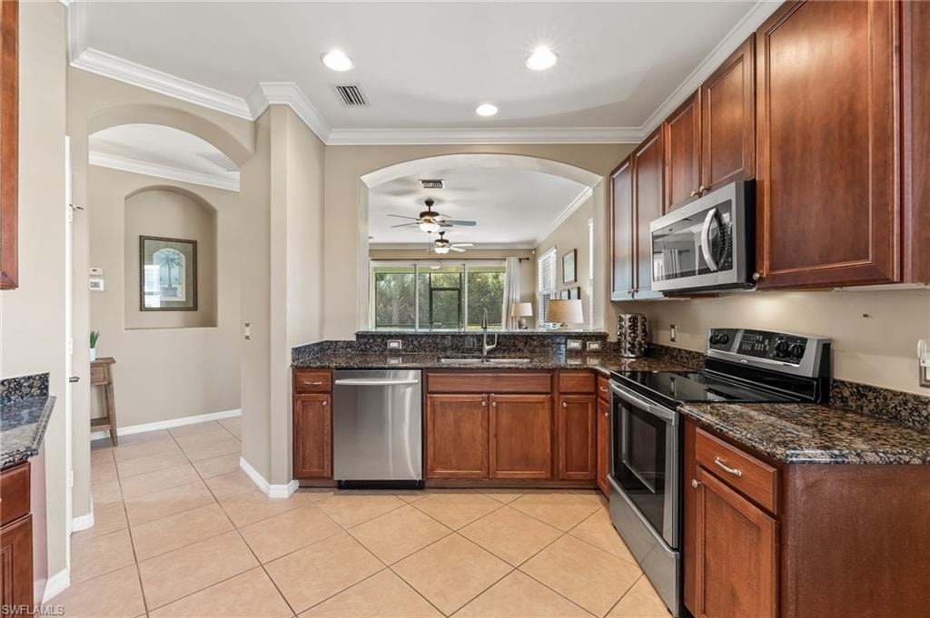 Kitchen featuring stainless steel appliances, a ceiling fan, dark stone counters, arched walkways, and light tile patterned flooring