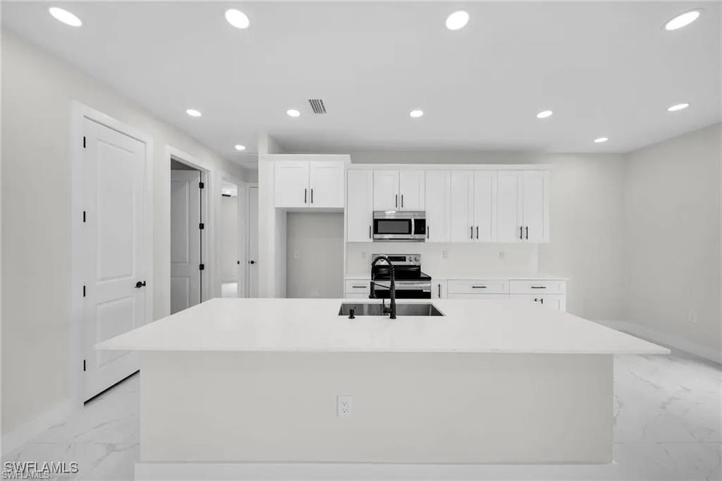 Kitchen with recessed lighting, white cabinets, stainless steel appliances, a center island with sink, and light marble finish flooring