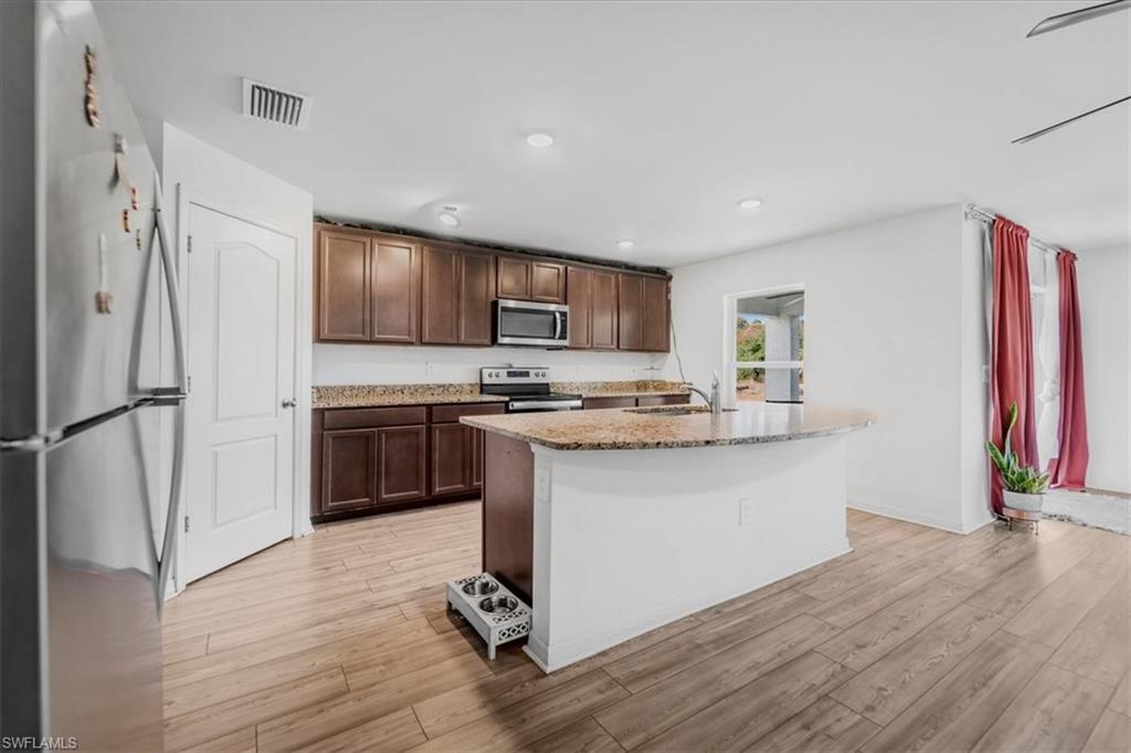 Kitchen with stainless steel appliances, light stone counters, a center island with sink, light wood-type flooring, and dark wood finish cabinetry