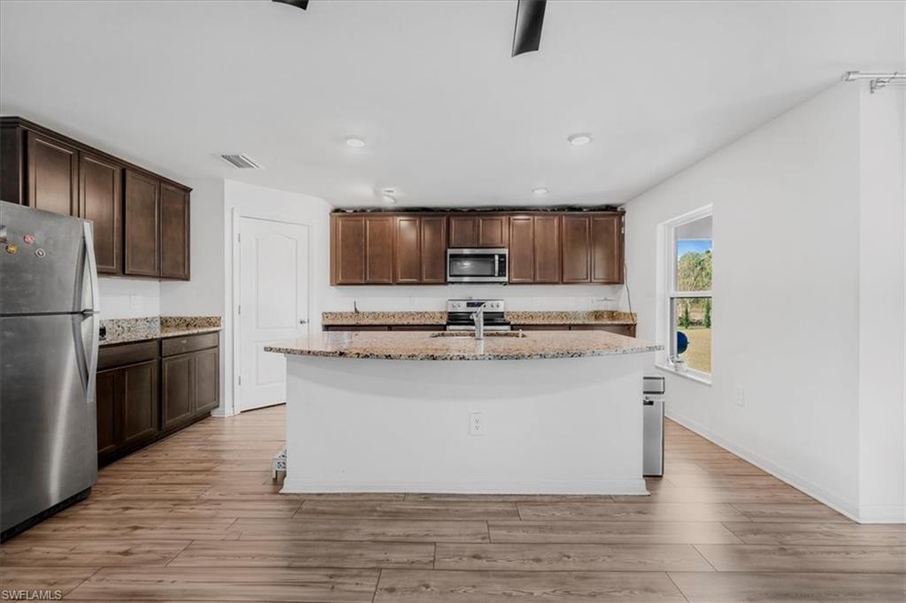 Kitchen featuring light stone counters, stainless steel appliances, dark wood finish cabinets, light wood-style flooring, and an island with sink