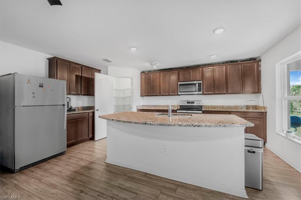 Kitchen featuring stainless steel appliances, light wood finished floors, a kitchen island with sink, dark wood finish cabinets, and light stone counters