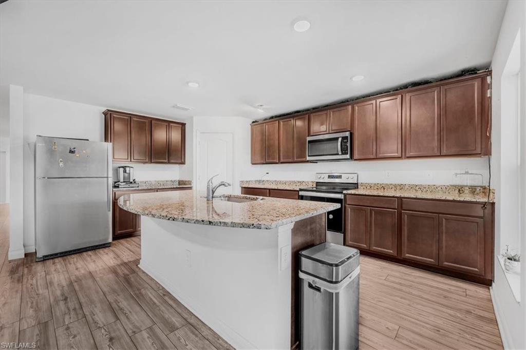 Kitchen featuring stainless steel appliances, light stone counters, dark wood finish cabinetry, light wood finished floors, and a center island with sink