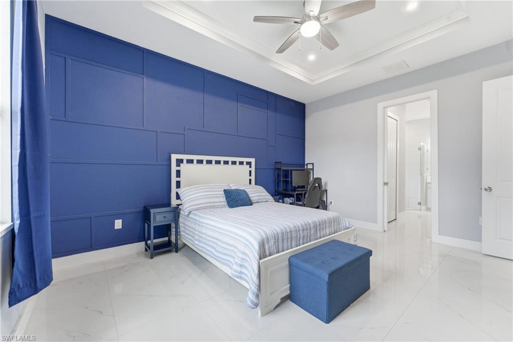 Bedroom featuring a raised ceiling, an accent wall, a decorative wall, light marble finish floors, and ceiling fan